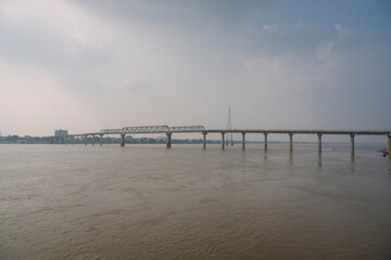 A long bridge spans across the Ganges River in Varanasi, India, under a cloudy sky, connecting both sides of the city.