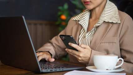 Businesswoman working on laptop and checking financial information on smartphone in hand in minimalist office with hot cocoa drink and documents on desk.