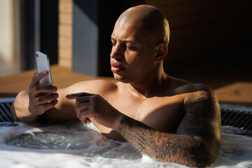 Tattooed african american man using smartphone in hot tub with hydro massage