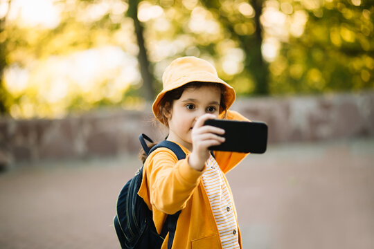 Cute little girl taking selfie with mobile phone looking at camera in a park. Kid blogger recording video vlog. - Powered by Adobe