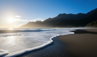 Fototapeta premium A scenic view of a tranquil beach with the sun rising behind misty mountains, casting light on the shore.