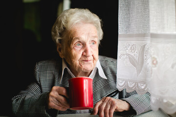 An elderly woman in a business suit sits at a table, holding a red mug, exuding wisdom and grace in...