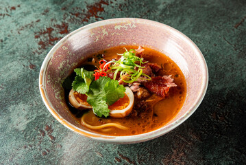A beautifully presented bowl of ramen soup with sliced meats, boiled eggs, and fresh herbs, resting on a textured green surface, highlighting vibrant colors and appetizing textures.