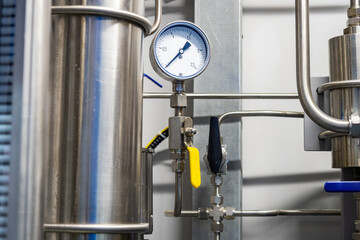 A stainless steel pressure gauge is mounted on an industrial piping system, with yellow and black control valves used for fluid regulation and monitoring