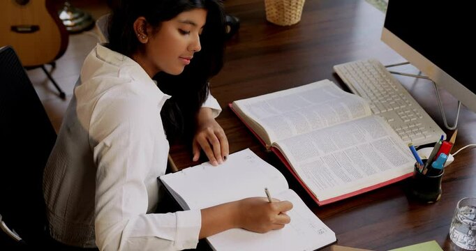 Indian Asian teenage girl studying at home, writing notes from a book, completing assignments or homework in a cozy lavish home, focused on education, exam preparation, and self-study in airy space
