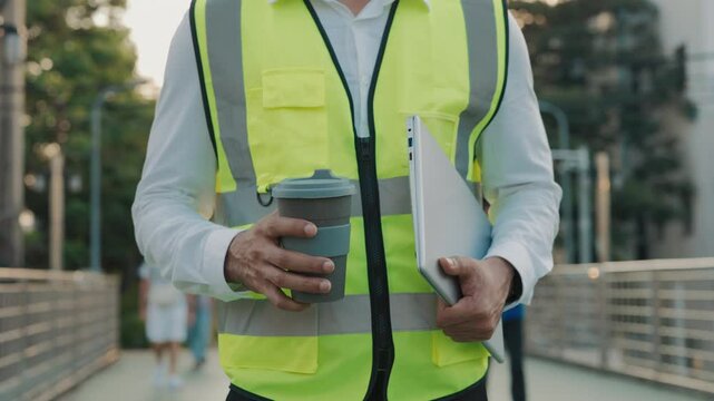 Man builder with laptop and coffee walks along footbridge against green park closeup. Experienced contractor wearing safety vest carries computer and cup to workplace outdoors