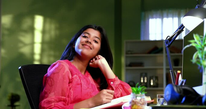 Indian Asian studious teenage beautiful girl sitting at a study table in a cozy lavish home, showing a confident success gesture while studying, writing, demonstrating focus, determination, excellence