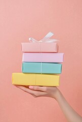 Hand holding a stack of four pastel colored gift boxes wrapped with string or ribbon on a peach background