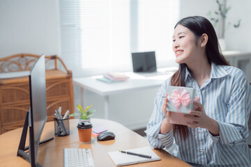 Young businesswoman smiling while holding a beautifully wrapped gift, engaging in a video call from her bright home office, radiating happiness and cheerfulness in her workspace