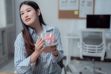 Young Asian office worker sitting at her desk, holding a gift box adorned with a pink bow, feeling a mix of curiosity and excitement about the surprise waiting inside
