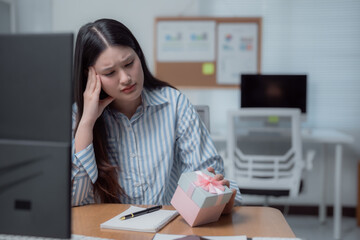 Asian office worker sitting at her desk, feeling sad and disappointed while holding an unwanted...