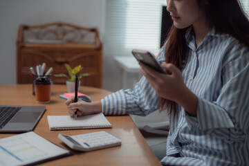 Young Asian businesswoman working from home, sitting at wooden table and taking notes on notepad while consulting smartphone, with laptop and calculator nearby