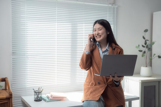 Smiling Asian businesswoman multitasking at her desk, using a laptop while communicating on her mobile phone, enjoying the view from a modern office window