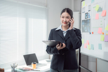 Young Asian businesswoman wearing suit is talking on phone and holding clipboard while standing near whiteboard with sticky notes and graphs in office
