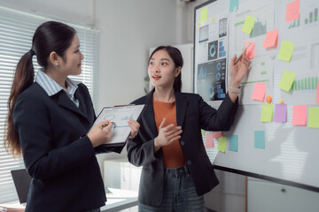 Two Asian businesswomen engaging in strategy discussions while utilizing a whiteboard filled with colorful sticky notes and printed graphs in a bright, modern office environment