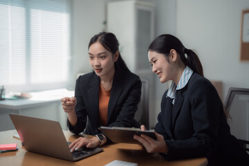 Two asian businesswomen are smiling and working together using a laptop in the office, one is pointing at the laptop screen while the other is holding a clipboard