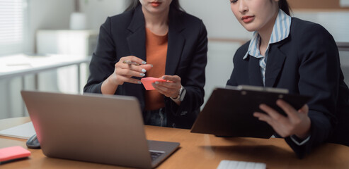 Two businesswomen collaborating in a modern office, utilizing sticky notes and a laptop, while one reads from a clipboard, focused on planning and strategizing for their project