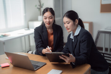 Fototapeta premium Two asian businesswomen analyzing financial data using laptop and clipboard discussing project strategy during a meeting in modern office, brainstorming and sharing ideas
