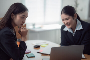 Two Asian businesswomen collaborating in a modern office, working together on a laptop while discussing and sharing innovative ideas for their project and strategies