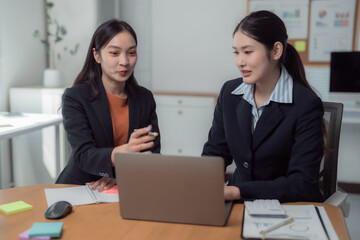 Two asian businesswomen are discussing business strategy, working and analyzing using laptop computer with financial document data graph and chart at office desk