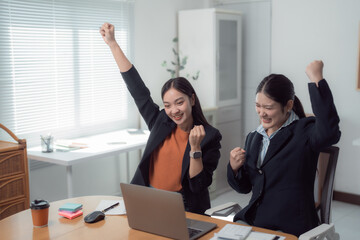 Two asian businesswomen are raising their arms and smiling, celebrating success while sitting at a desk with a laptop, calculator, and sticky notes in a modern office