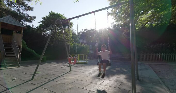 Young boy swinging at a playground, basking in the sunlight, with rays shining through trees, surrounded by play structures, enjoying a carefree and joyful moment outdoors