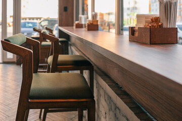 Cozy modern cafe interior with wooden chairs and high counter along the window. Warm sunlight creates a welcoming atmosphere. Minimalist design with natural wood and neutral tones