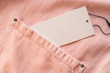 Blank white tag on pink denim jacket with metal buttons. Fashion label for branding or price. Close-up of clothing texture, pocket detail, soft pastel color. Mockup for text or logo