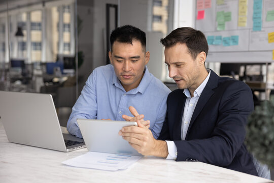 Two diverse company managers working on project together, using digital gadgets for teamwork, corporate discussion, watching online web product presentation on tablet computer