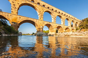 Fototapeta premium Pont du Gard, ancient Roman aqueduct bridge, Provence late afternoon golden light on arches mirroring in river, Gorges du Gardon, natural landscape designated Biosphere Reserve by UNESCO, France