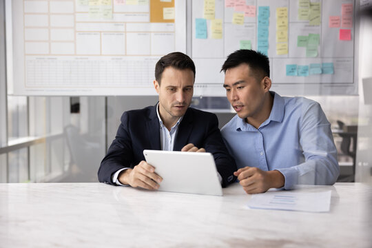 Serious Latin businessman consulting male Asian business expert, showing online content, project report on tablet to advisor, asking for advice, expertise, professional consultation