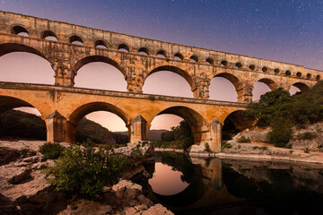 Pont du Gard, ancient Roman aqueduct bridge, colorful distant sunrise, under Provence starry night sky, Gorges du Gardon, natural landscape designated Biosphere Reserve by UNESCO, France
