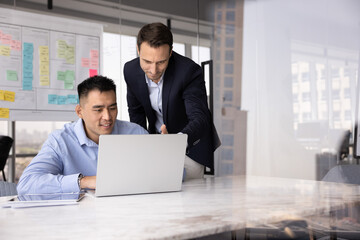 Male Caucasian project leader giving advice, help, instructions to Asian employee. Two colleagues talking at computer, sitting and standing at workplace, pointing and looking at screen