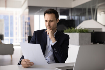 Pensive concerned Hispanic business legal expert man reading financial document, checking contract, examining agreement text at workplace, touching chin, thinking