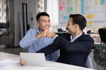 Two happy multiethnic businessmen finding successful decision on brainstorming meeting at tablet computer, making fist bump hand gesture, getting agreement, smiling, laughing