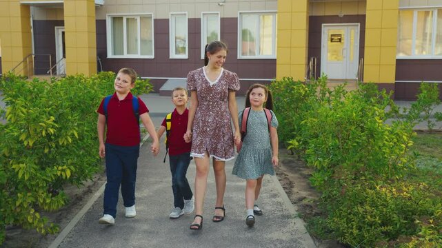 Mother leading children from school, female mother holding children hand, happy family, young nanny with children, schoolchildren with backpacks walking with aunt by the hand, little boys girl walking