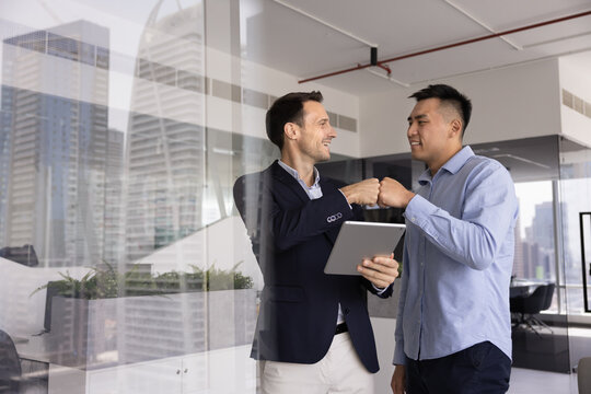 Two positive male business colleagues enjoying partnership, friendship, giving fist bump due to successful effective teamwork, using tablet computer, standing in office, talking, laughing