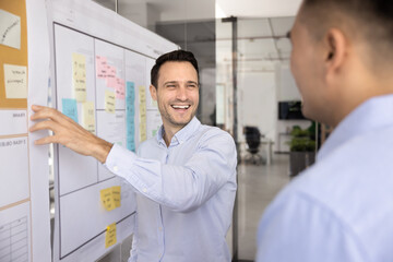Fototapeta premium Cheerful male Hispanic project manager presenting startup teamwork plan on scrum boards pointing at whiteboard to business partner, talking to Asian colleague man, smiling, laughing