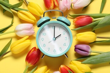 Spring time. Alarm clock and beautiful tulips on yellow background, flat lay
