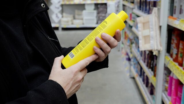 Person carefully reading label of bright yellow shampoo bottle while comparing products in store aisle, selecting personal care item with attention to details