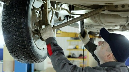 Skilled mechanic wearing protective gear examining vehicle undercarriage, using flashlight to check...