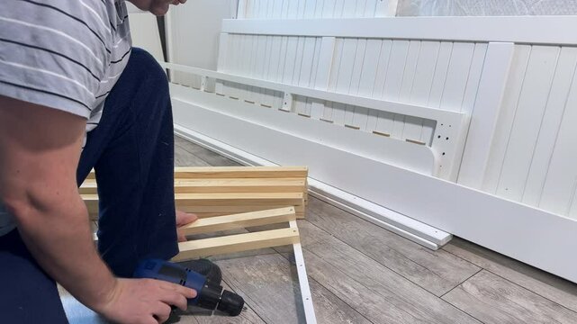 A man assembles the base of a couch. He fastens wooden slats with screws. Using a screwdriver. Side Profile Shot .
