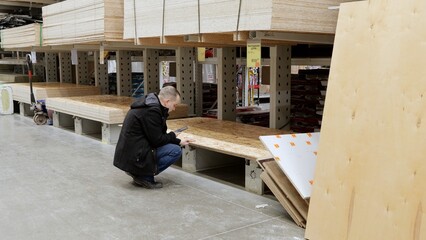 Fototapeta premium Customer crouching, browsing smartphone while selecting wood planks among stacked construction panels and materials inside hardware store warehouse