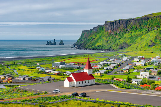 Panoramic view of basalt stacks Reynisdrangar and yellow meadow flowers at Vik town with Myrdal church, Iceland, at summer sunny day and blue sky.