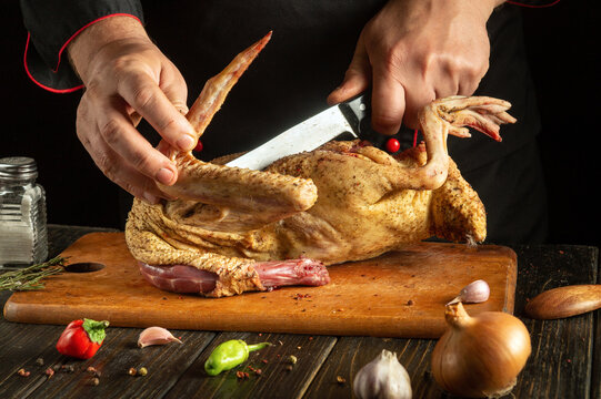 A skilled chef uses a sharp knife to cut raw poultry on a wooden cutting board. Fresh herbs, garlic, and chilies surround the area, showcasing a vibrant culinary setting