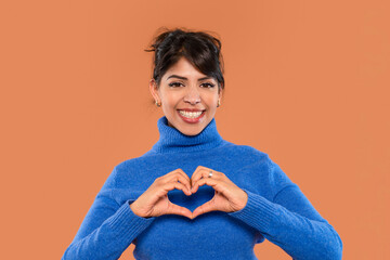 Smiling woman in blue sweater forming heart shape with her hands against warm brown background in...
