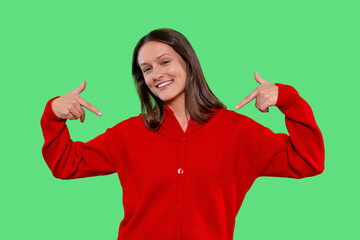 Young woman in red cardigan smiling and pointing at yourself on green background during cheerful...