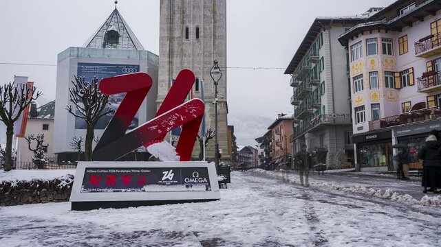 Timelapse of a snowy town square in Cortina d'Ampezzo, Italy, featuring the Olympic countdown clock, bell tower, alpine buildings, and pedestrian activity.