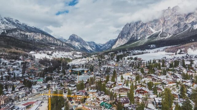 Aerial view of Cortina d'Ampezzo, showing snow covered rooftops, a yellow crane, preparing for the Winter Olympics 2026, dense forests, and dramatic Dolomite mountain peaks.