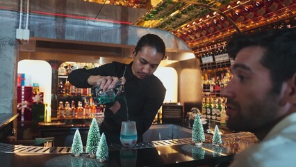 Bartender pouring mixed blue lagoon cocktail liqueur to cooling glass while guest talking friends waiting drink, decorated pine light christmas theme making special festive tasty in couple. Vinosity.